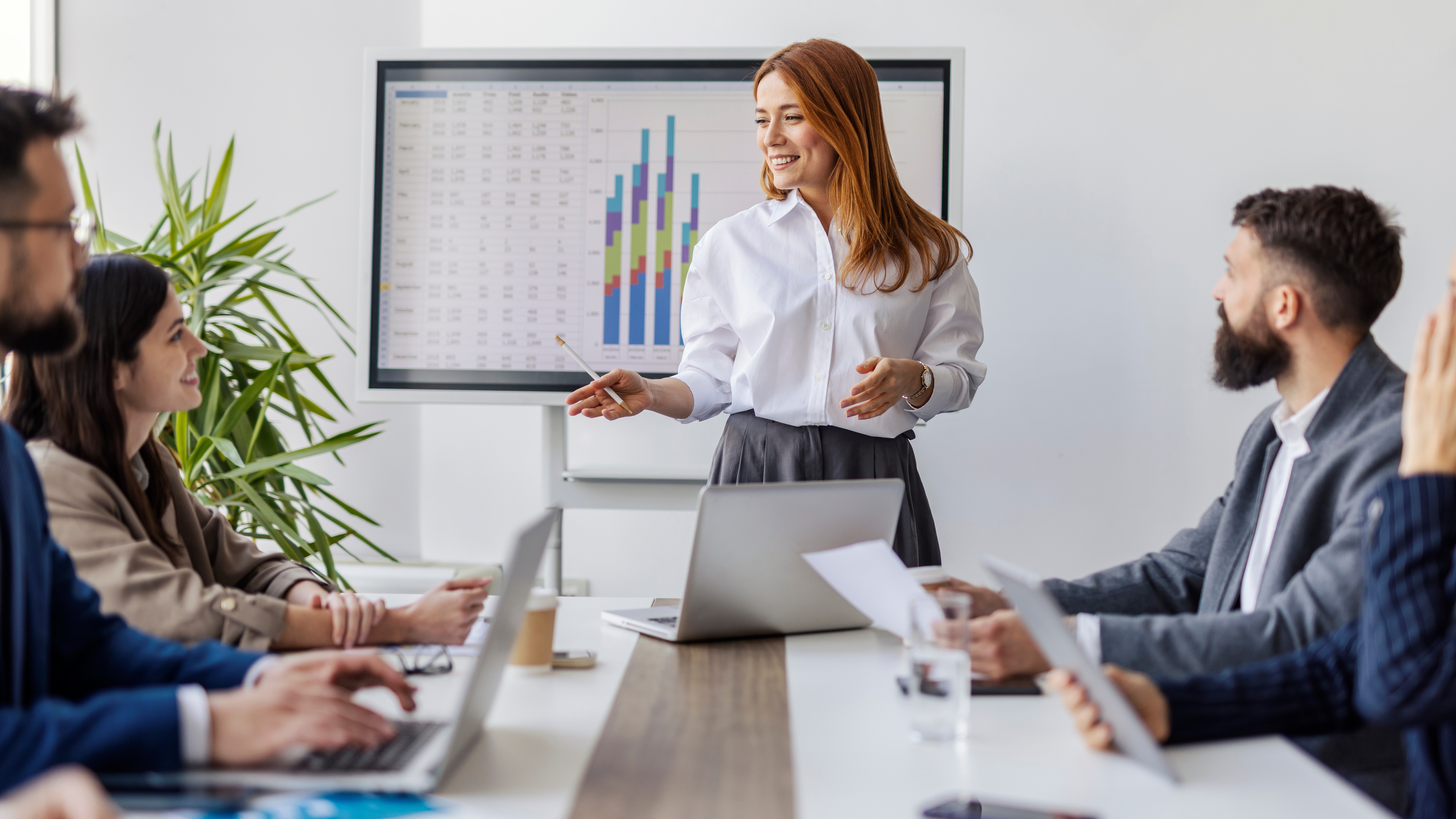 Portrait of female executive standing near conference table at boardroom with her leadership team and explaining financial report.
