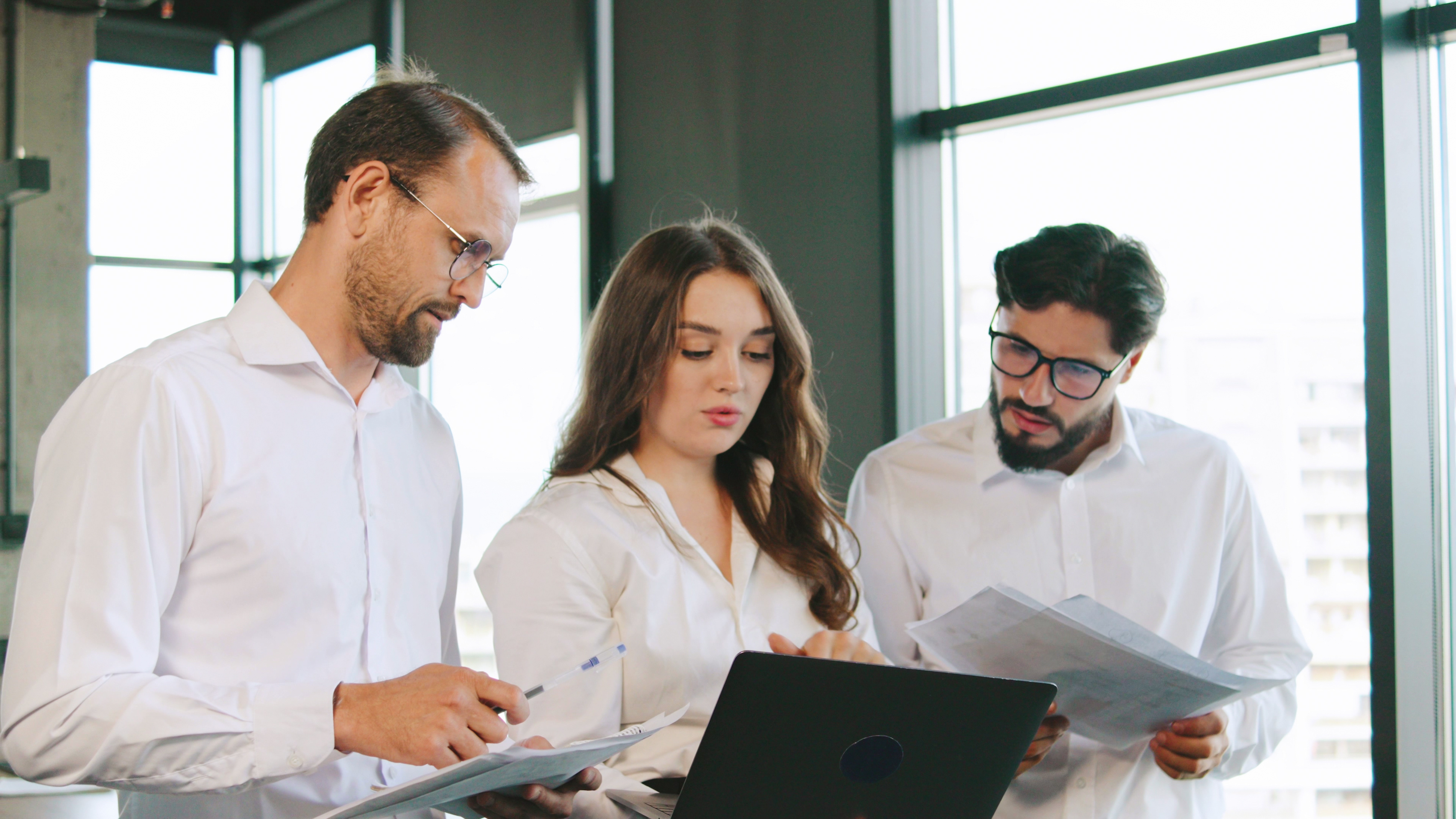 Three professionals review project documents and data on a laptop while collaborating in a bright office setting, sharing ideas and insights.
