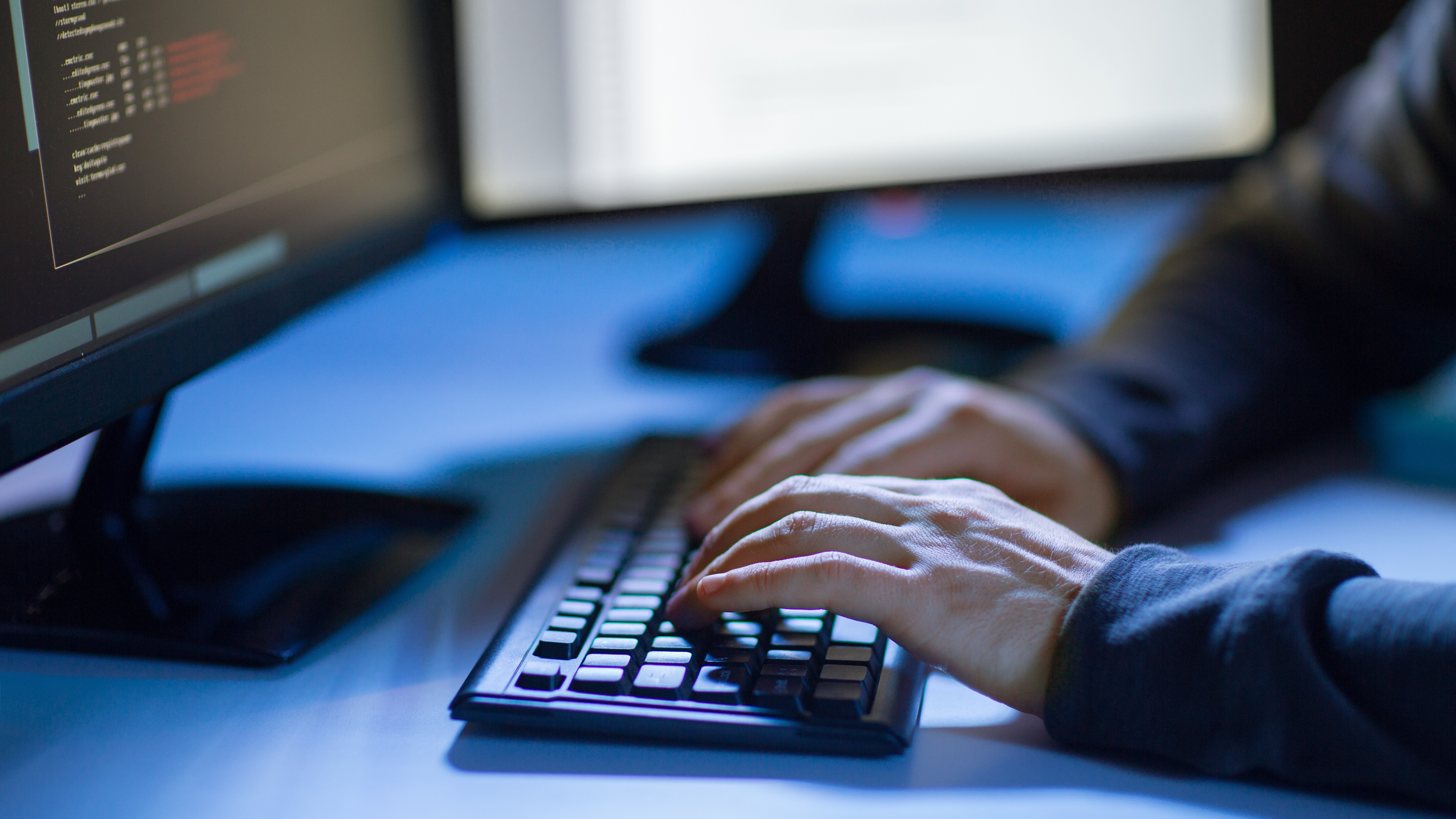 Hacker sitting in a dark room typing on a keyboard, illuminated by computer screens displaying code.