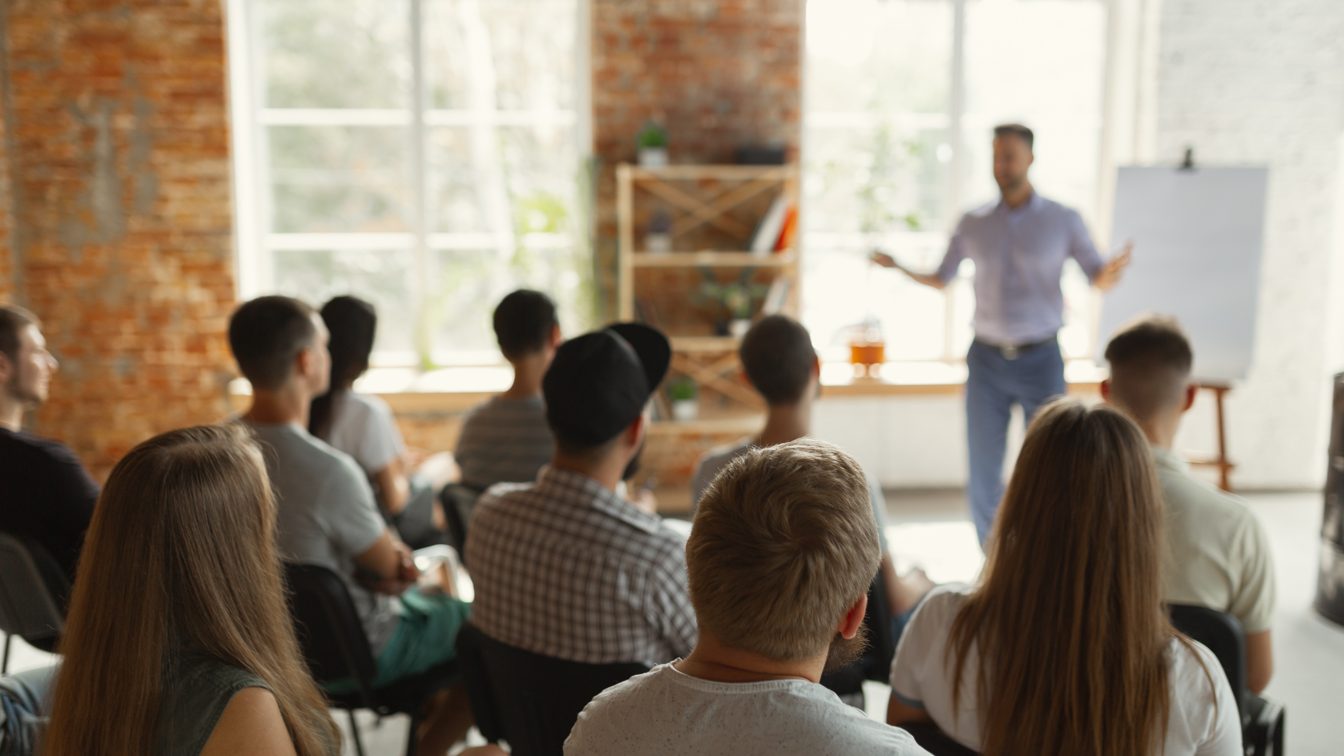 Male speaker giving presentation in hall at university workshop. Audience or conference hall. Rear view of unrecognized participants in audience. Scientific conference event, training. Education See Less